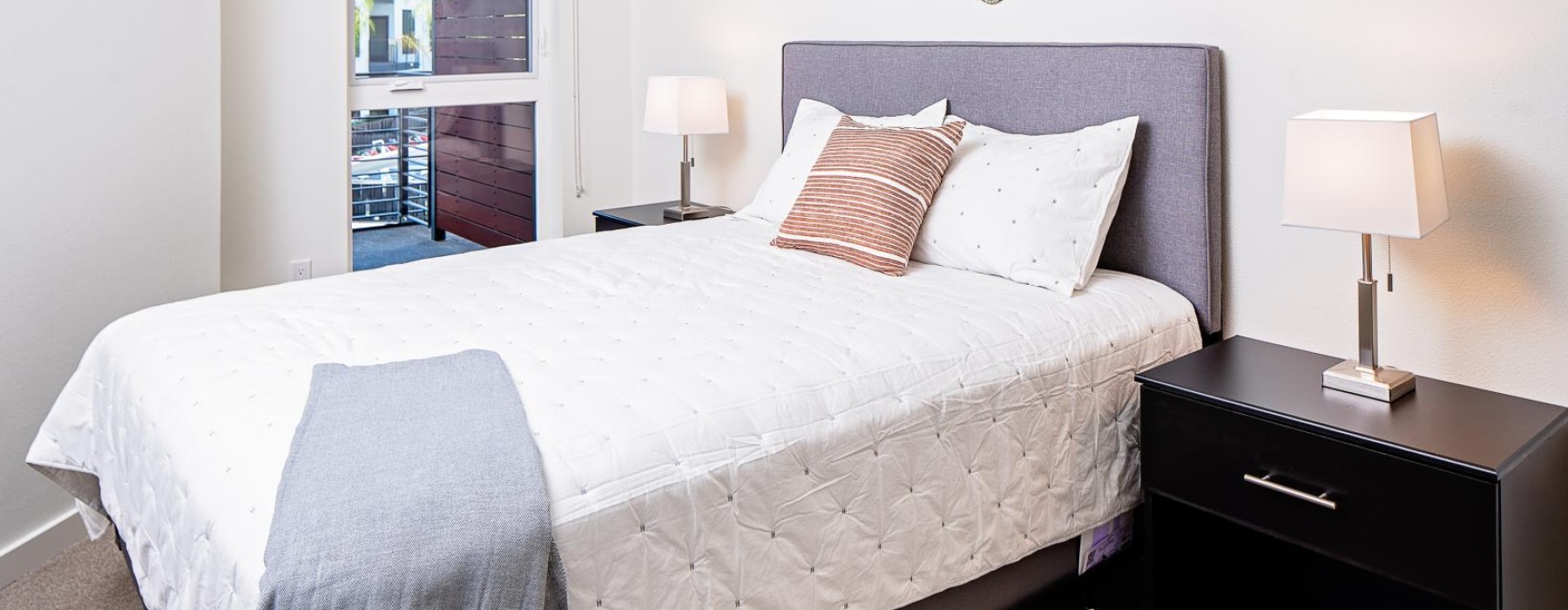 A modern bedroom with a white-clothed bed, grey headboard, and two bedside lamps next to a window.