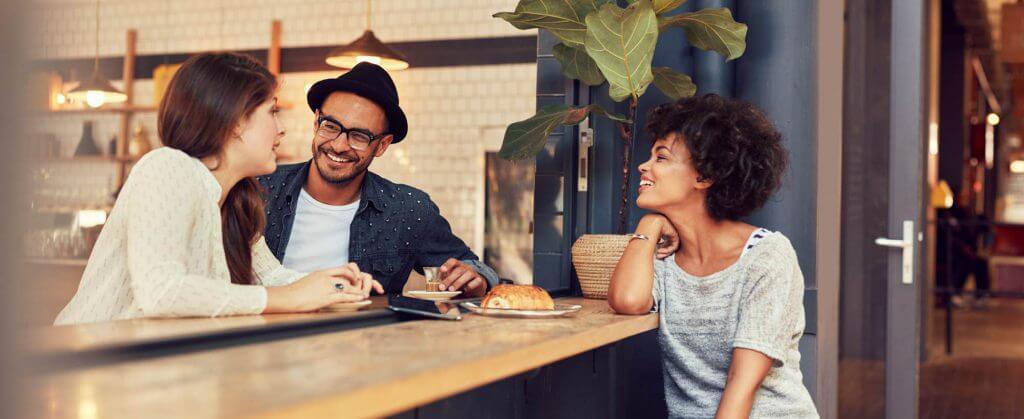 A group of three friends enjoying a conversation at a stylish café counter, with drinks and pastries on the table.